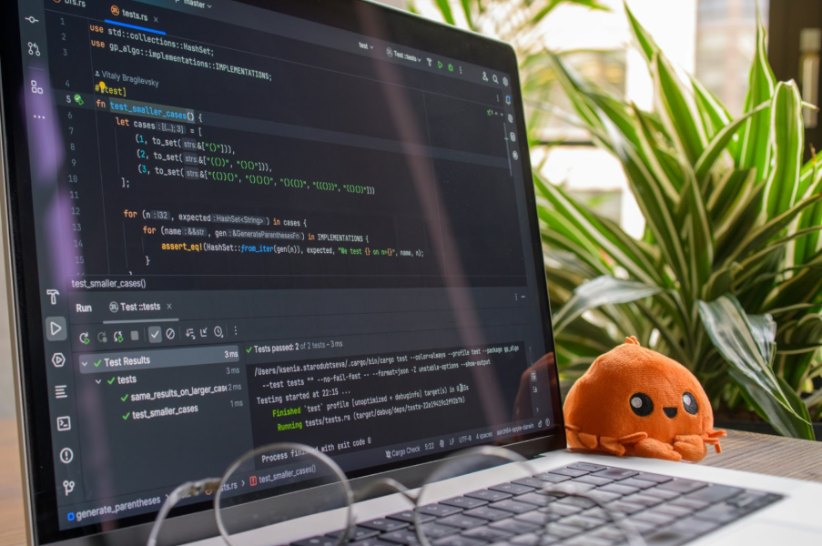 A close-up of a laptop screen displaying a code editor, alongside a pair of glasses, a small plant, and an orange plush toy.